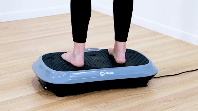 Woman standing barefoot on a vibration plate in a bright living room.